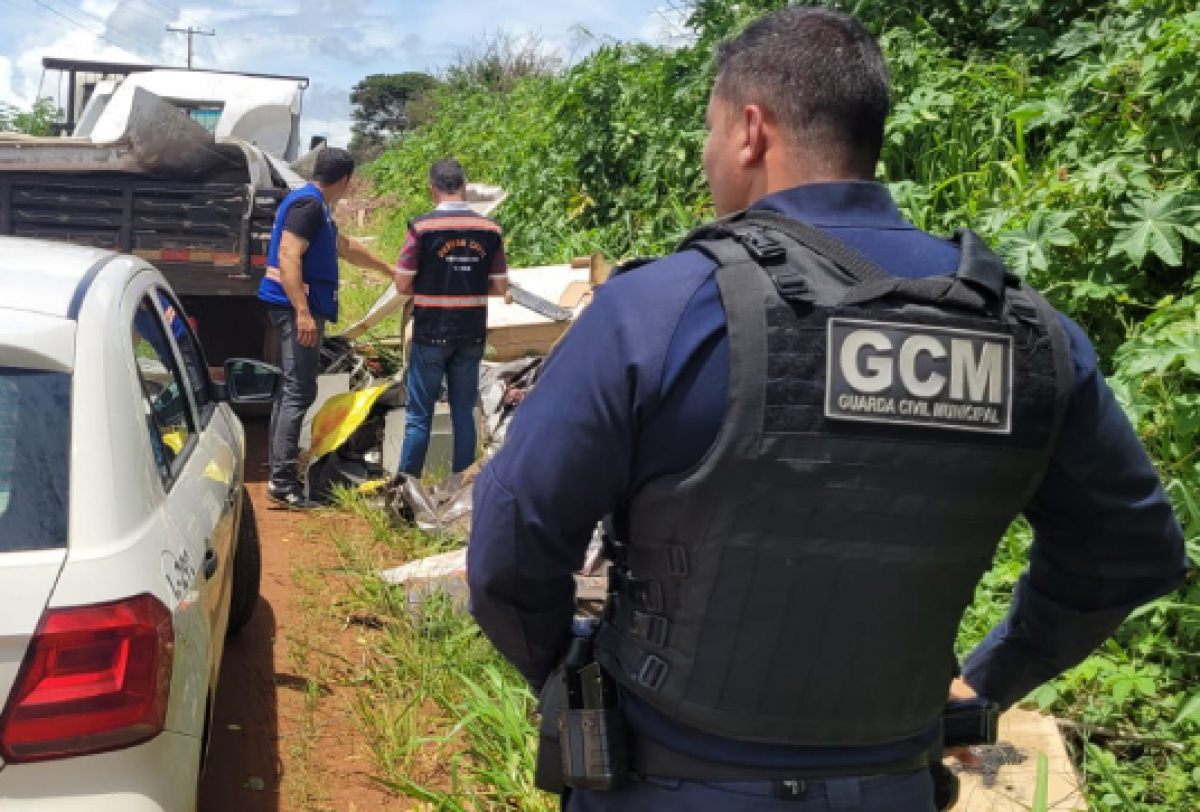 Um guarda civil municipal inspeciona um monte de destroços ao lado de uma estrada de terra, com outros indivíduos e veículos ao redor.